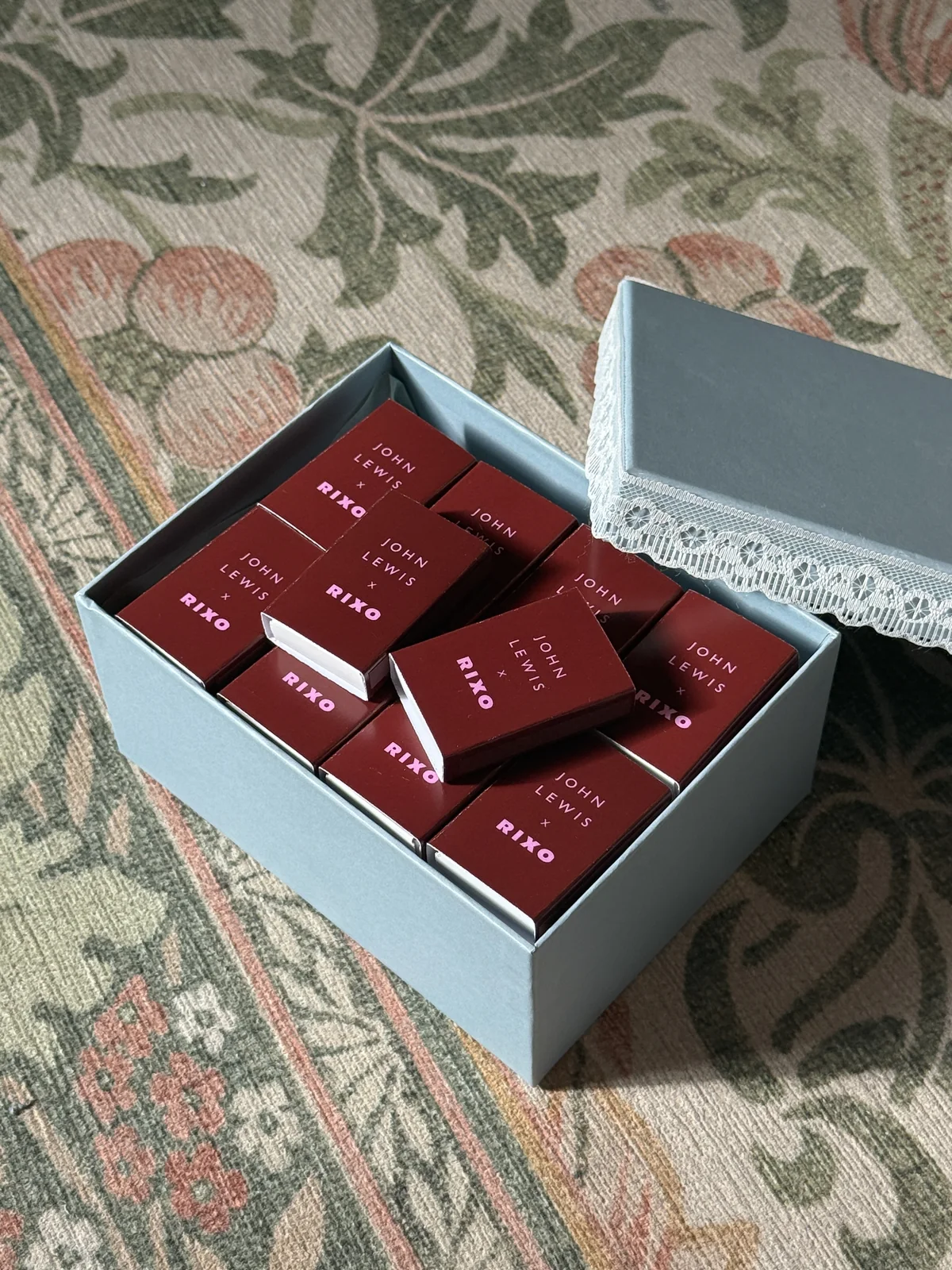 Deep red John Lewis × RIXO matchboxes lettered in pink, in a pale blue box with a lace-edged lid, on a patterned carpet in sunlight