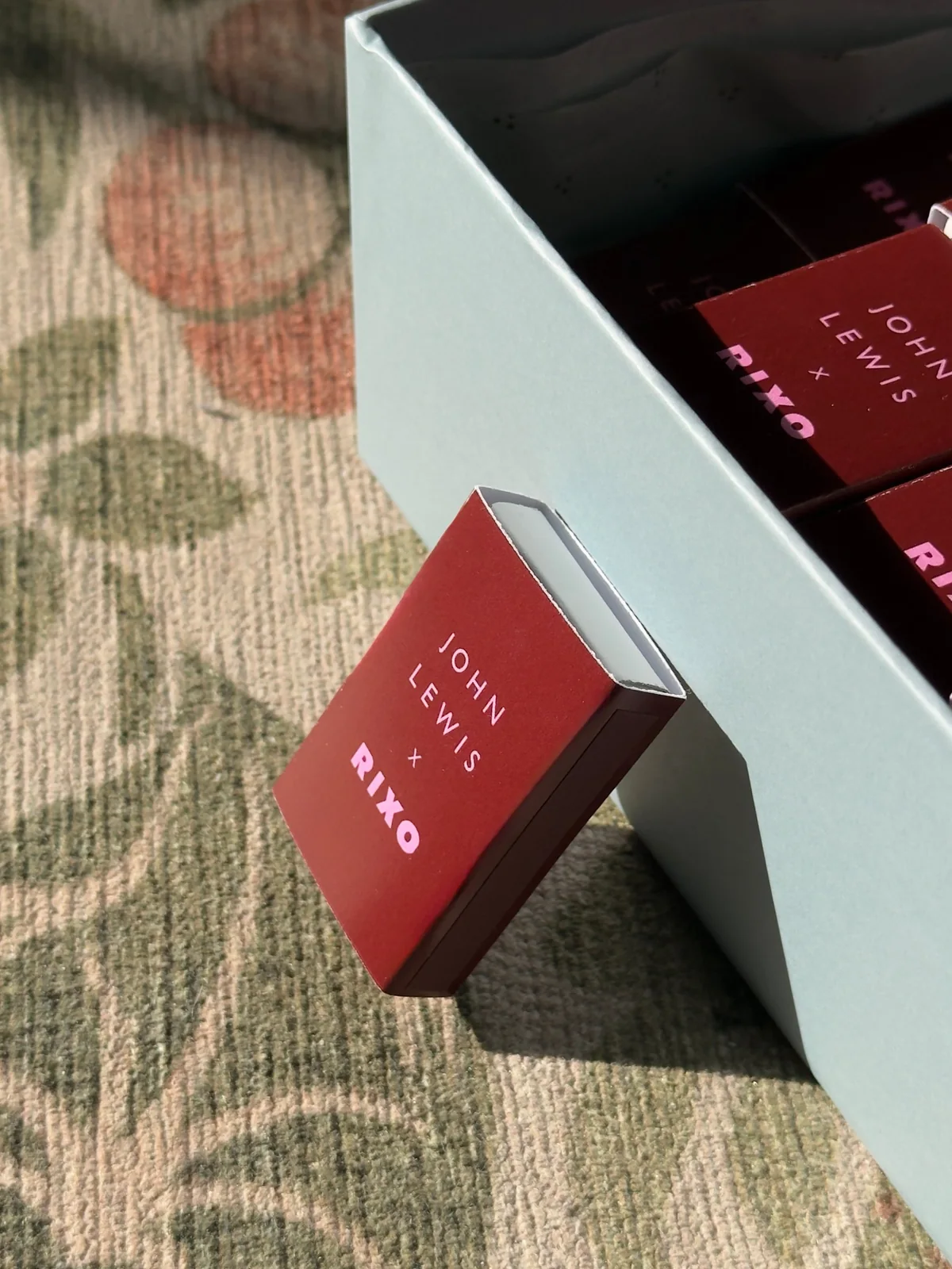 A single John Lewis × RIXO matchbox resting against the edge of its blue box