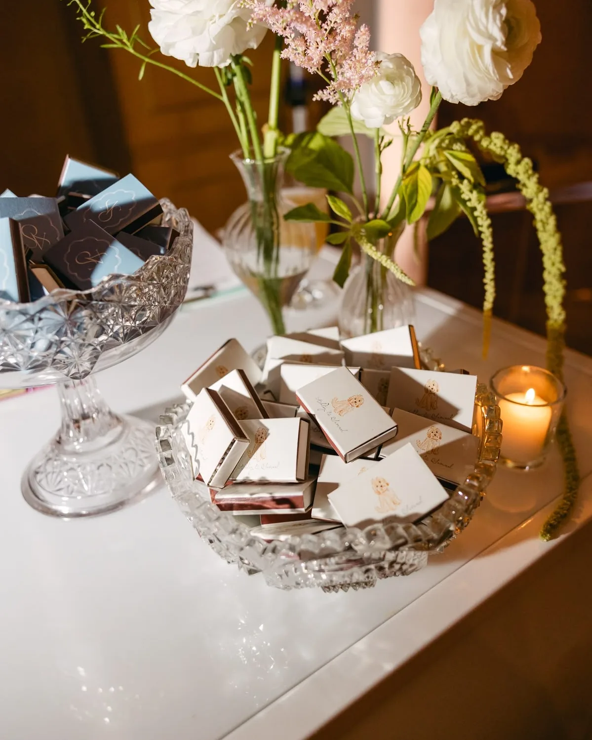 Illustrated matchboxes in a crystal bowl beside white ranunculus and a candle