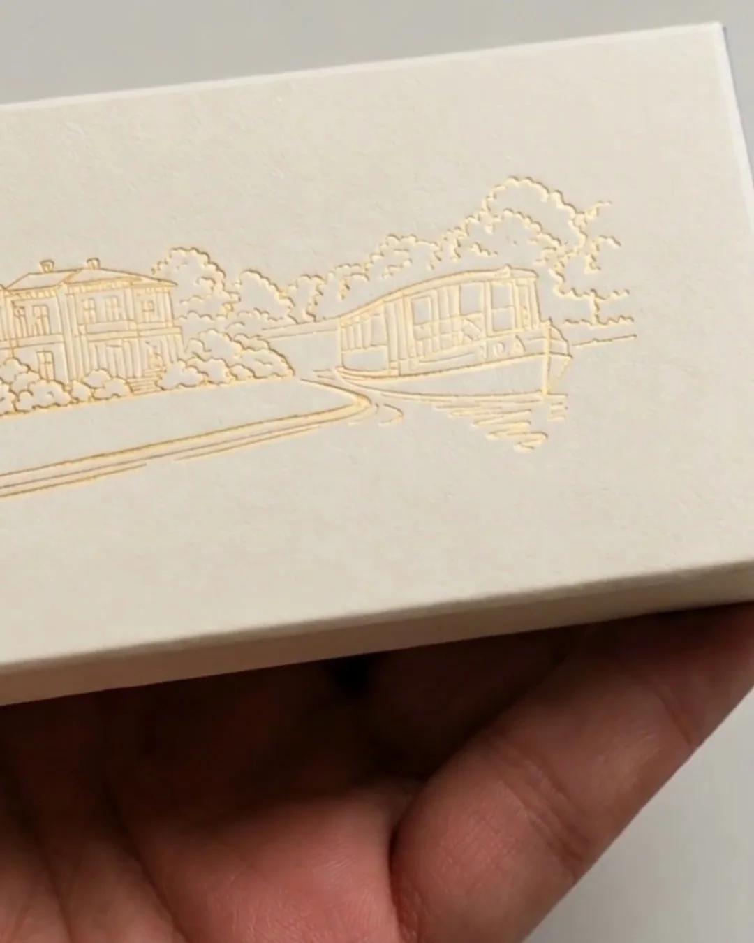 A cream box with a gold embossed drawing of a house and garden, held in the hand