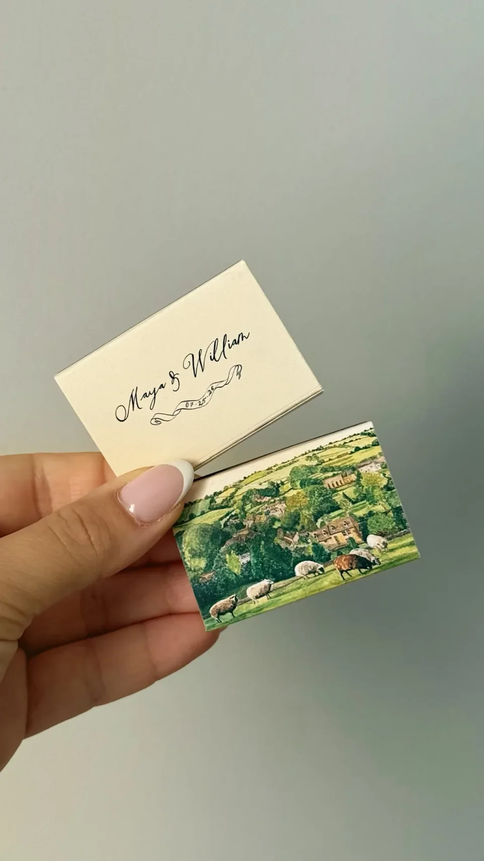 A cream matchbox with two names in script and one with a painted countryside of fields and sheep