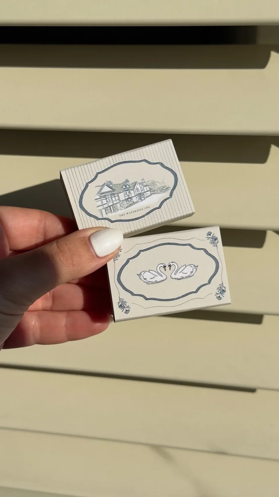 Cream matchboxes with an inn drawn in a blue frame and two white swans