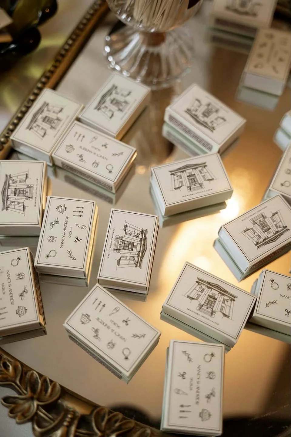 Ivory wedding matchboxes drawn with a townhouse and small icons, scattered on a gilded mirror tray