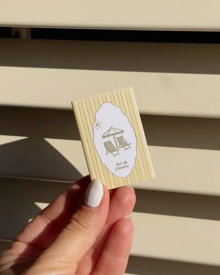 A yellow striped matchbox with two drawn deckchairs and the words July 26, Croatia, held in the sun